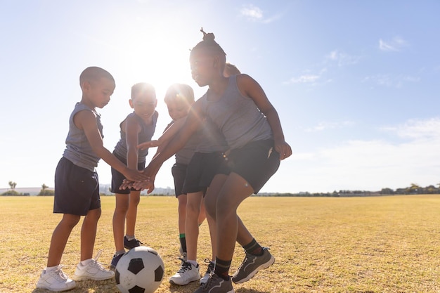 Des écoliers élémentaires multiraciaux empilant les mains sur un ballon de football sur le terrain contre le ciel aux beaux jours