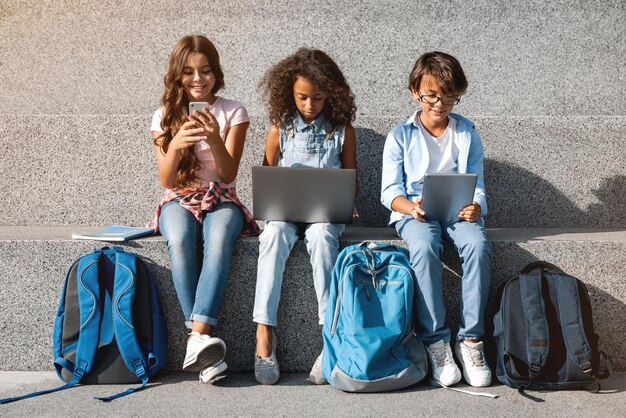 Des écoliers souriants assis avec des appareils numériques dans la rue