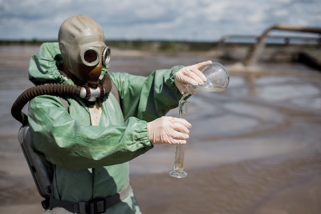 Un écologiste masculin dans une combinaison de protection verte et un masque à gaz prélève un échantillon d'eau Le scientifique fait une étude toxicologique Post apocalypse