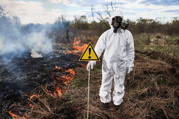 Photo un écologiste pompier éteignant un incendie sur le terrain un homme en combinaison de protection contre les rayonnements et un masque à gaz près de l'herbe brûlante avec de la fumée tenant un panneau d'avertissement avec un signe d'exclamation concept de catastrophe naturelle