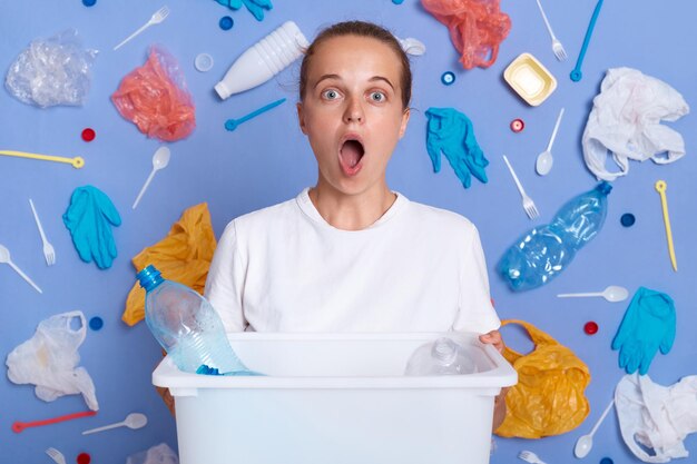 Une écologiste stupéfaite garde la bouche grande ouverte impressionnée par un gros problème environnemental ramasse des déchets réutilisables et tient une boîte avec des bouteilles en plastique contre les ordures du mur bleu autour