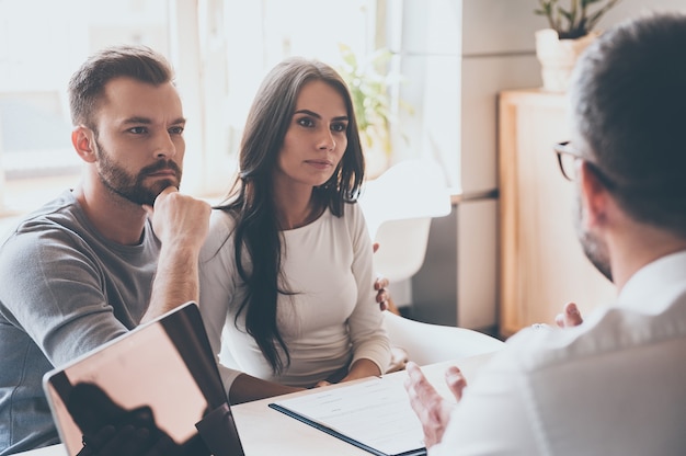 A l'écoute du conseiller financier. Jeune couple concentré se liant les uns aux autres et écoutant un homme assis devant eux au bureau du bureau