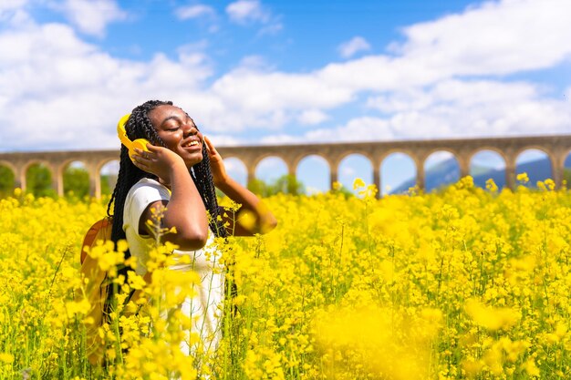 Écouter de la musique dans des écouteurs jaunes une fille ethnique noire avec des tresses un voyageur dans un champ de fleurs jaunes