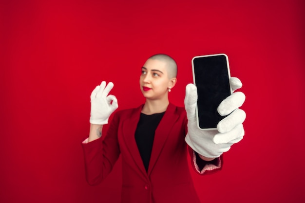 L'écran du téléphone vide. Portrait de jeune femme chauve caucasienne isolée sur mur rouge. Beau modèle féminin en gants. Émotions humaines, expression faciale, ventes, concept publicitaire. Culture bizarre.