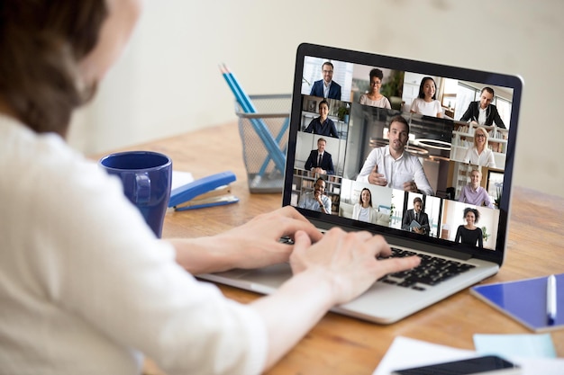 Sur l'écran de l'ordinateur portable, la webcam affiche divers hommes d'affaires d'âge et d'ethnie qui participent à un appel vidéo de groupe, vue sur l'épaule de la femme, assis au bureau travaillant à domicile Concept d'application moderne de communication à distance