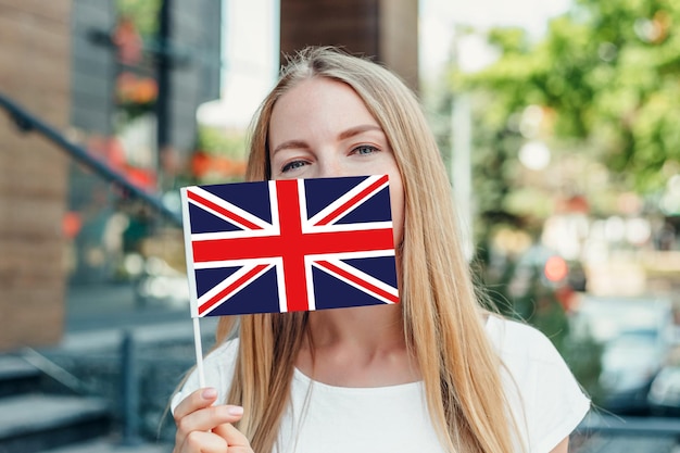 Éducation à L'étranger. Une étudiante Se Couvre Le Visage D'un Petit Drapeau Britannique De L'angleterre Britannique Et Se Tient Près De L'université Dans La Rue