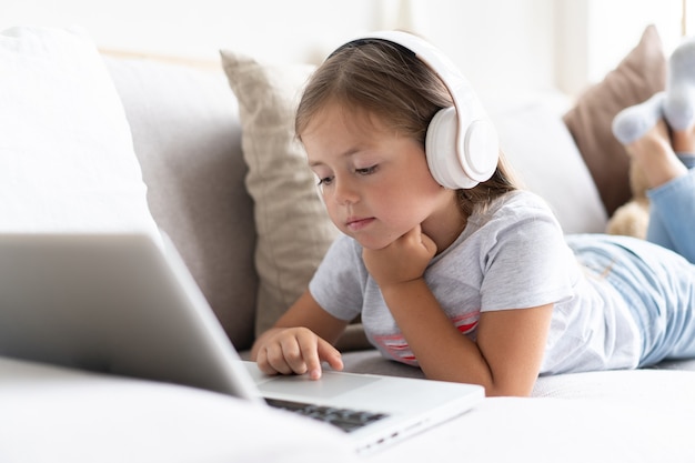 L'éducation en ligne des enfants. Fille douce avec des écouteurs à la recherche d'un ordinateur portable de conférence de professeur de cours vidéo assis sur le canapé à la maison.