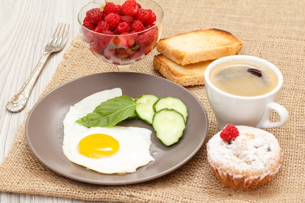 egguf frit, concombre frais et basilic, muffin, tasse de café, toasts et bol en verre avec framboises.