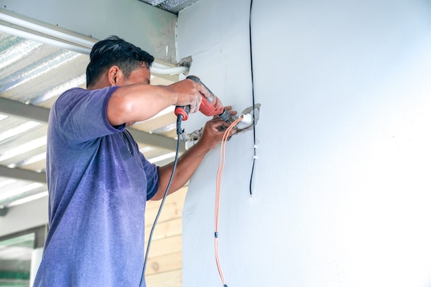 Un électricien asiatique tente de réparer l'interrupteur et le fil électrique sur le mur détruit.