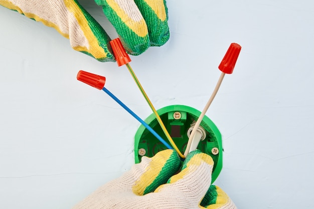 Un électricien en gants de protection met des sertissages sur des fils serrés à l'intérieur d'un boîtier électrique rond pour prise.