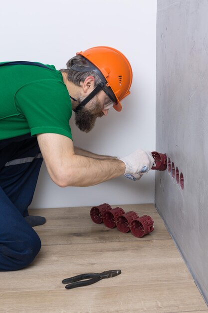 Un électricien installe des prises dans l'appartement Un gars dans un casque orange et une salopette fait de l'électricité dans la maison