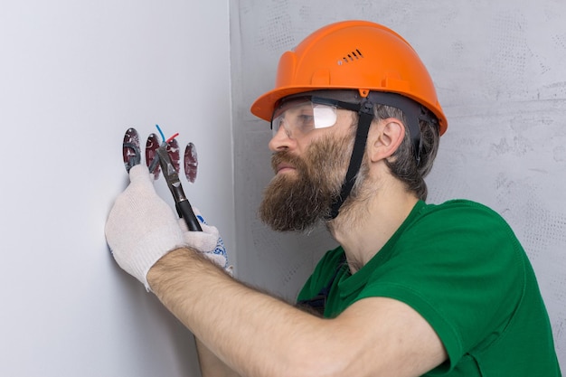 Un électricien installe des prises dans l'appartement Un gars dans un casque orange et une salopette fait de l'électricité dans la maison