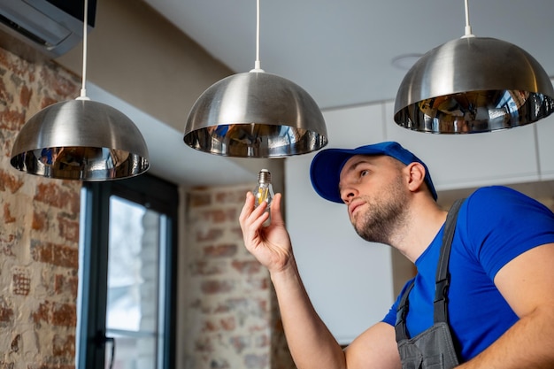 Un électricien mâle change les ampoules de la lampe Un jeune homme en salopette effectue des travaux techniques sur l'électrification des locaux