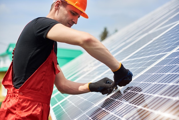 Un électricien en t-shirt noir et gants installe une cellule solaire pour économiser de l'énergie contre le ciel bleu