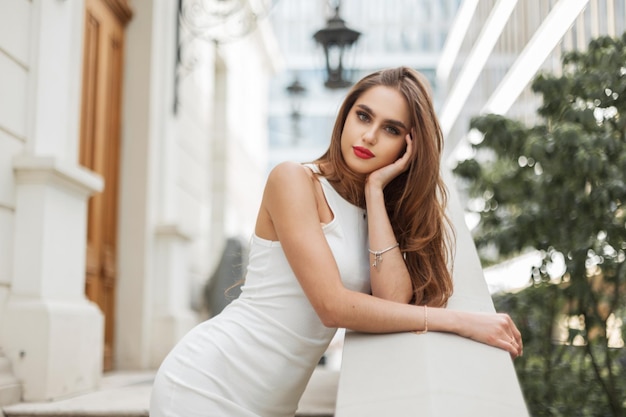 Elegant belle jeune fille de la mode avec des lèvres rouges dans une robe blanche élégante se tient et pose près d'un bâtiment vintage dans la ville