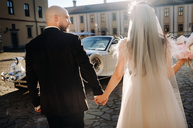 Un élégant couple de mariage Dans la cour du château se promène en se tenant la main près d'une voiture rétro