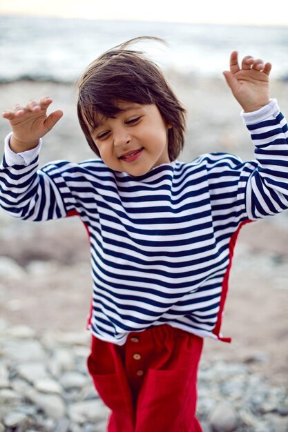 Photo Élégant garçon enfant dans un gilet s'asseoir sur une plage de galets au bord de la mer en été