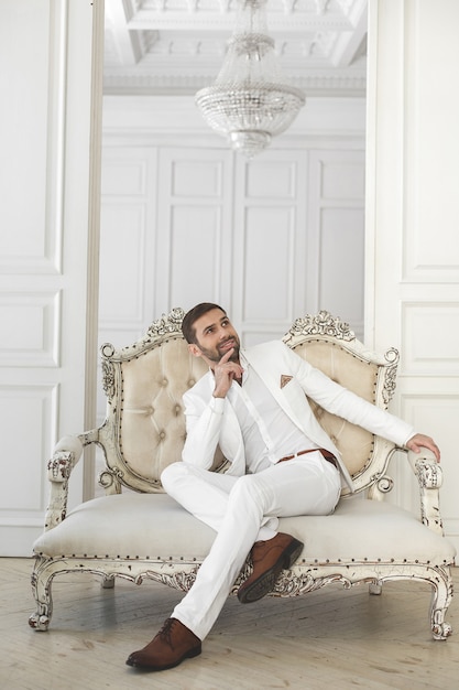 Photo Élégant jeune bel homme avec une barbe dans un costume classique blanc. l'intérieur de l'hôtel. studio de photographie. canapé. la fenêtre. chaise. lustre