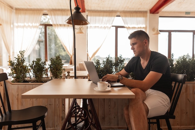 Photo Élégant jeune pigiste dans des vêtements à la mode est assis à une table dans un café vintage et travaille sur un ordinateur portable moderne à l'heure du déjeuner.