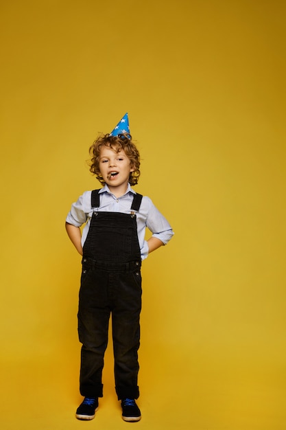 Élégant Petit Garçon En Salopette Et Bonnet D'anniversaire Posant Sur Le Fond Jaune, Isolé. Mode Enfantine