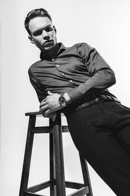 Un élégant portrait noir et blanc d'un homme en chemise posant sur un cyclorama blanc avec une chaise