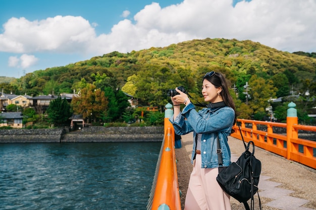 Photo Élégant voyageur asiatique prenant une photo sur le pont avec une belle montagne en arrière-plan. dame lentille homme photographier la rivière propre à kyoto. voyage de vacances de printemps au japon.