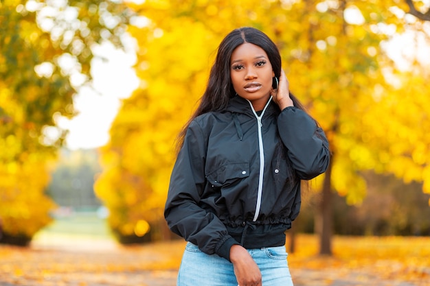 Élégante Belle Jeune Femme Afro-américaine Dans Une Veste à La Mode Avec Un Jean Se Promène Dans Un Parc Sur Fond De Feuillage D'automne Jaune