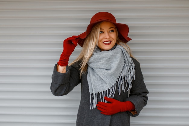 Photo Élégante belle jeune femme dans un chapeau à la mode avec une écharpe grise dans un manteau élégant avec des gants posant près d'un mur en bois