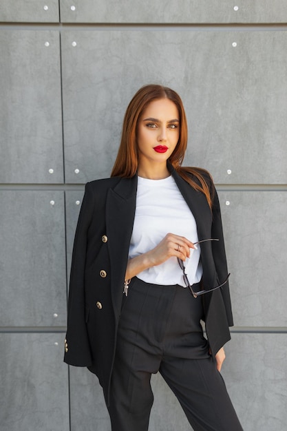 Photo elegante belle jeune femme décontractée avec des lèvres rouges dans des vêtements de mode avec un t-shirt blanc et un blazer noir avec des lunettes de soleil se tient près d'un mur gris dans la ville trendy jolie dame chic