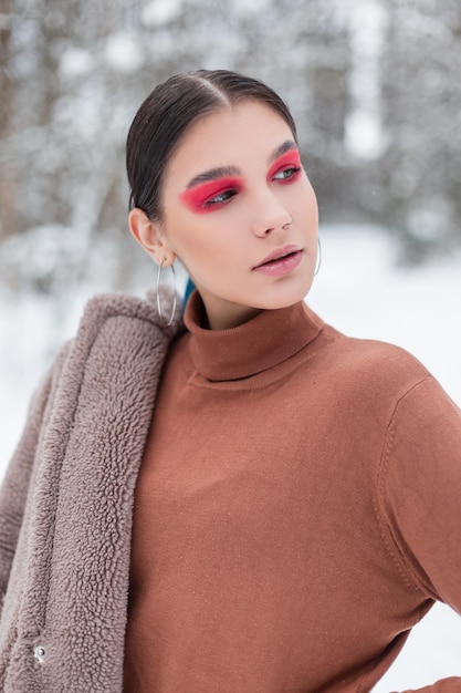 Élégante belle jeune femme de mode avec un maquillage lumineux et des ombres à paupières roses dans un pull et un manteau vintage à la mode posant dans une forêt d'hiver enneigée