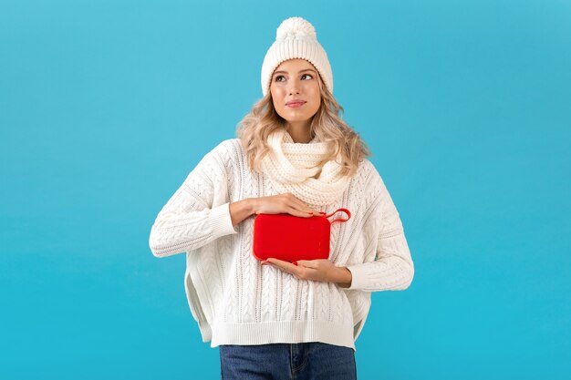 Élégante Blonde Souriante Belle Jeune Femme Tenant Haut-parleur Sans Fil écouter De La Musique Heureux Portant Chandail Blanc Et Bonnet Tricoté Style Hiver Posant Isolé Sur Mur Bleu