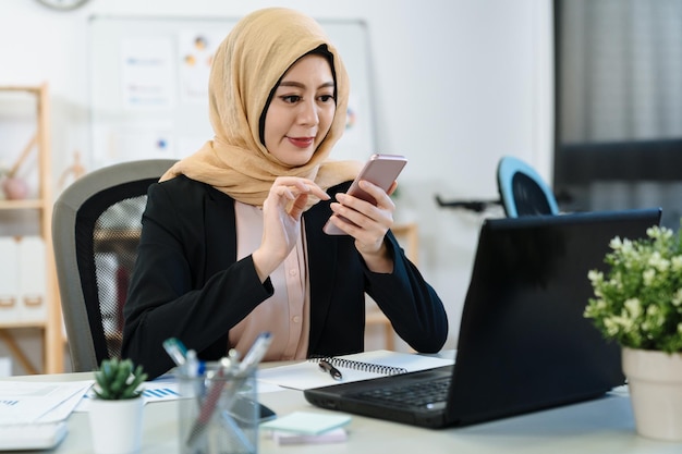 élégante femme d'affaires musulmane travaillant sur un ordinateur portable dans un bureau moderne. détendez-vous l'islam travailleuse avec foulard jaune souriant en utilisant un téléphone portable discutant en ligne pendant la pause-café en entreprise.