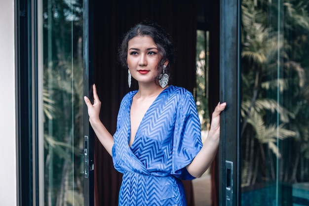 Une élégante femme asiatique vêtue d'une robe bleue à col bas ouvre des portes vitrées sur sa villa; concept de luxe exotique.