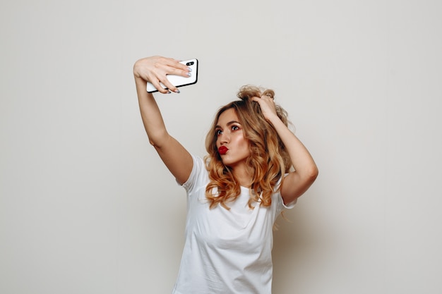 élégante femme blonde aux lèvres rouges prend selfie sur smartphone sur blanc.