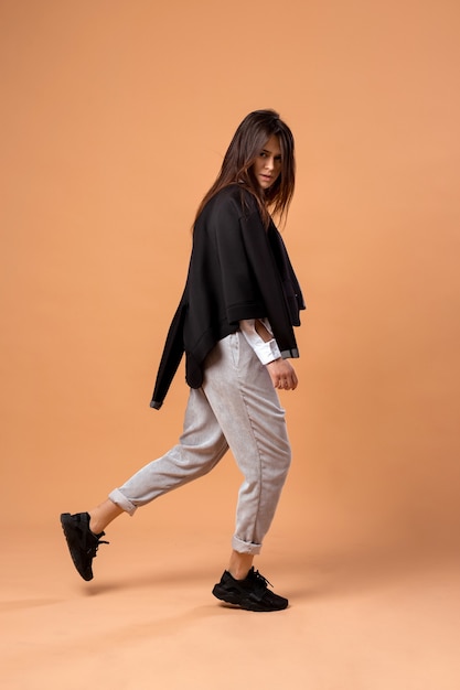 Une élégante fille aux cheveux noirs vêtue d'une chemise blanche, d'un pantalon gris, d'une veste noire et de baskets noires pose sur fond beige dans le studio.