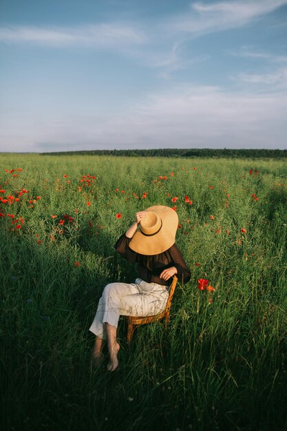 Photo Élégante fille élégante au chapeau assis sur une chaise rustique dans la prairie d'été avec des fleurs