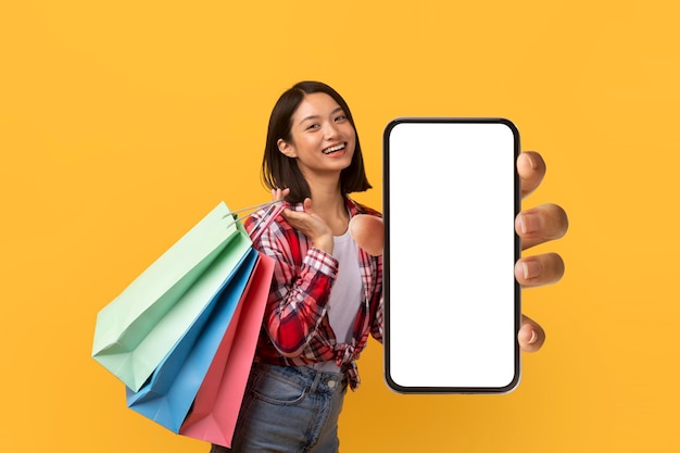 Élégante jeune femme asiatique posant avec des achats et montrant un téléphone portable avec un écran vide sur une maquette de fond jaune