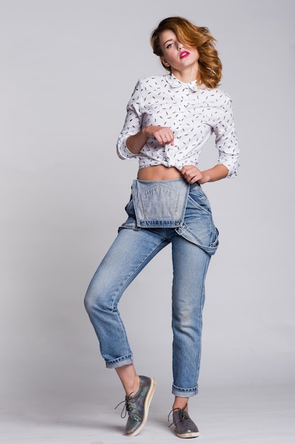 élégante jeune femme en combinaison denim jeans, chemise posant sur fond blanc