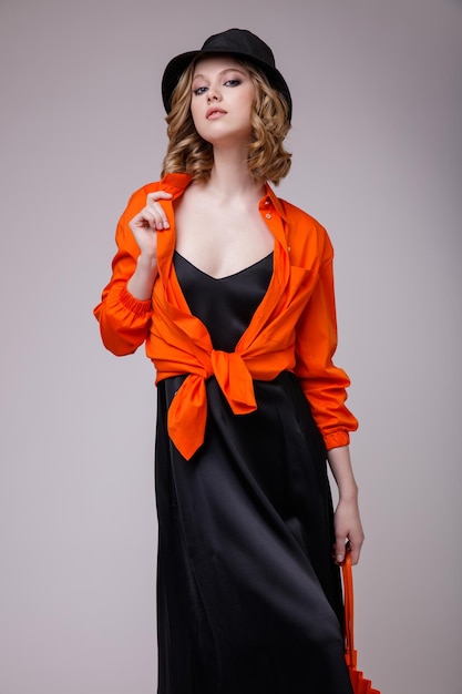 élégante jeune femme dans une robe noire chapeau chemise orange sac à main posant sur fond blanc