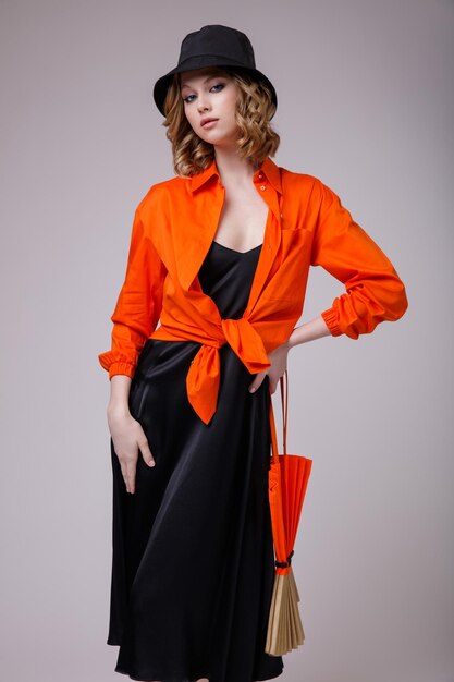 élégante jeune femme dans une robe noire chapeau chemise orange sac à main posant sur fond blanc