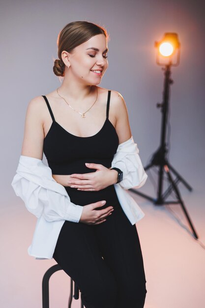 Une élégante jeune femme enceinte vêtue d'une chemise blanche et d'un pantalon noir est assise sur une chaise et serre son ventre sur un fond blanc Une jolie femme avec un maquillage professionnel et des vêtements décontractés