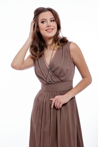 Élégante jeune femme en robe marron beige avec un collier décolleté posant sur fond blanc