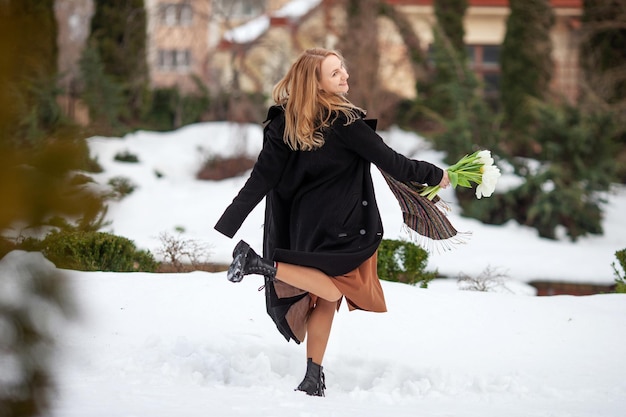 Élégante jeune femme souriante aux longs cheveux blonds en manteau noir dans la ville