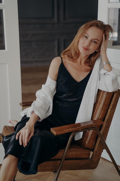 Élégante jolie jeune femme caucasienne vêtue d'une robe noire assise sur une chaise confortable se relaxant à la maison