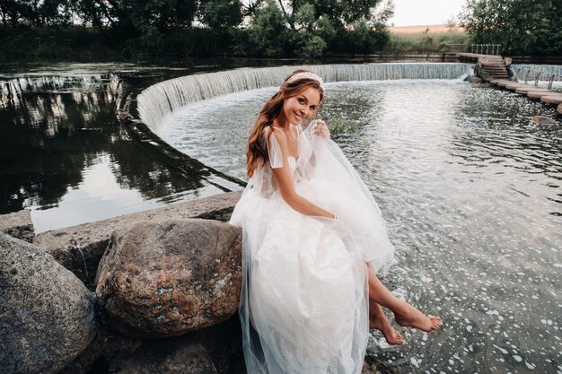 Une élégante mariée vêtue d'une robe blanche, de gants et de pieds nus est assise près d'une cascade dans le parc, profitant de la nature.Un modèle en robe de mariée et des gants dans un parc naturel.Biélorussie