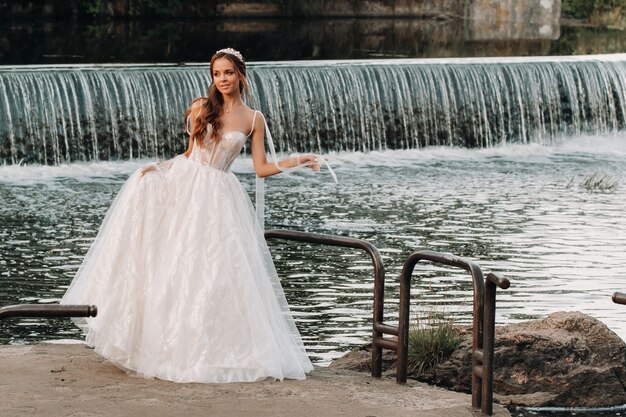 Une élégante mariée vêtue d'une robe blanche et de gants se tient près de la rivière dans le parc, profitant de la nature.Un modèle en robe de mariée et des gants dans un parc naturel.Biélorussie