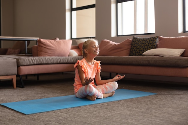 Une élémentaire caucasienne souriante médite alors qu'elle est assise sur un tapis de yoga près du canapé.