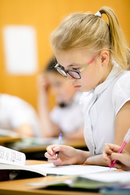 Photo Élève de l'école blonde mignonne avec des lunettes élégantes écrit en classe