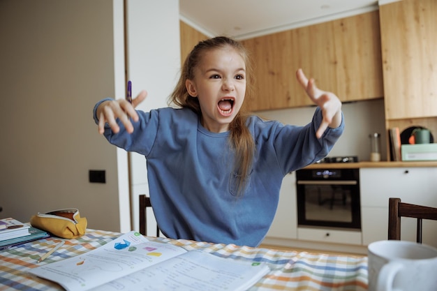 Photo une élève de première année rit émotionnellement, crie et se met en colère alors qu'elle fait ses devoirs à la maison dans la cuisine.