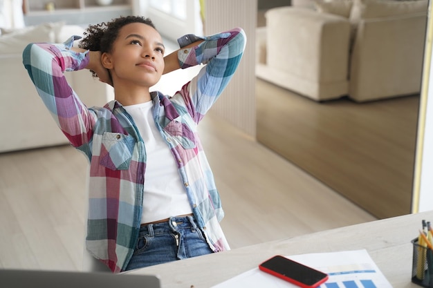 Une élève de race mixte décontractée aime faire une pause tout en apprenant à la maison en rêvant assis au bureau